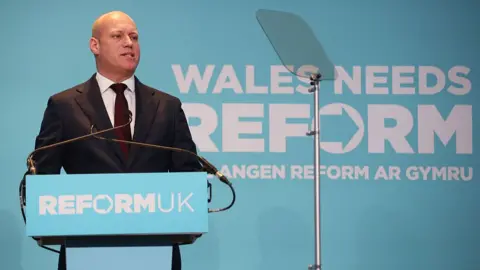 Getty Images A bald man stood at a Reform-branded podium with people sat in front of him