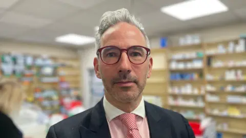 Ashley Cohen, wearing glasses, stands in a pharmacy wearing a white-collared shirt with red tie.