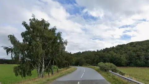 Picture of the B851, with trees on either side of the road and a blue sky with white clouds.