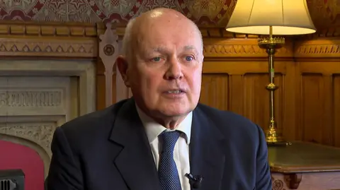 Sir Iain Duncan Smith sitting in front of a fireplace in a wood-panelled room, with a lampshade behind him