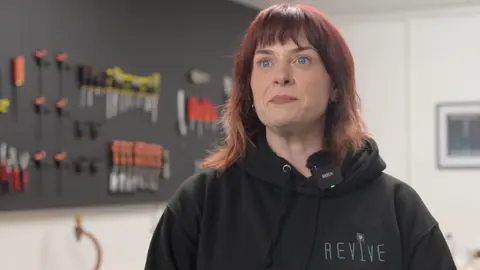 Mark McAlindon/BBC Rachel Stewart looking to the left of the camera with pursed lips. She had red hair with a full fringe and is wearing a black hoodie with the Revive logo on it. Behind her are various tools hanging on a wall, but they are out of focus.