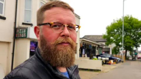 Paul Moseley/BBC Liam Calvert is standing in a street in Norwich and looking at the camera. He is wearing glasses.