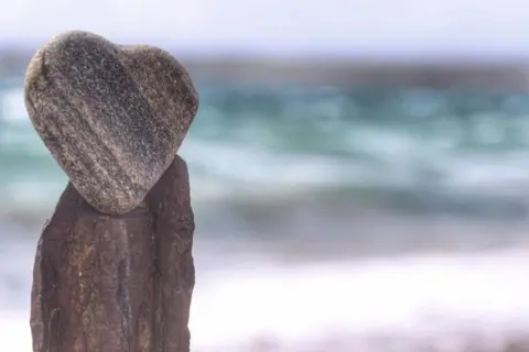 John Dyer A small "heart shaped" stone perched on top of another stone with rough waters out of focus in the background