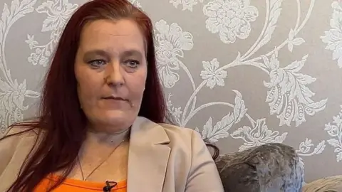 BBC Rebecca Ramsay with long brown hair wearing a biege blazer and orange top sitting on a velvet chair with floral patterned wallpaper in the background.