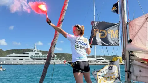 Lucy Tulloch A woman son standing on a sailboat, holding a lit red flare high in one hand. The flare emits bright red light and smoke that trails upward into the sky. She is wearing a white athletic shirt covered in various sponsor logos and dark shorts and has a harness or safety belt attached around the waist. The sailboat appears to be docked or sailing in calm blue water within a marina or coastal area. In the background, there are green hills and a large, white luxury yacht anchored nearby. Several other sailboats and masts are also visible behind the main subject. A black flag with gold lettering is attached to the sailboat's rigging and flutters in the wind. The boat’s deck and sails are partially visible, showing ropes, hardware, and additional sponsor logos.