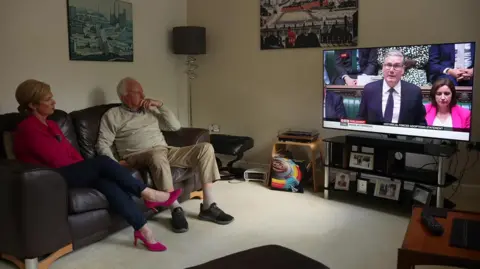 Steve Hindley and a woman in a pink top sitting on a black sofa as they watch Sir Keir Starmer in the House of Commons on TV. The living room has a black lamp in the corner and artwork on the walls.