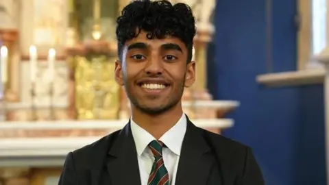 Shaun Whitmore/BBC Alfie is a teenage boy dressed in a black school blazer and white shirt. He is standing inside a church with an altar behind him.