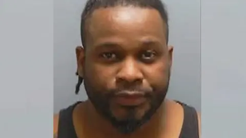 Bruno Sala has a peaked, dark beard and short, dark hair. He wears a black vest in a police custody photo.