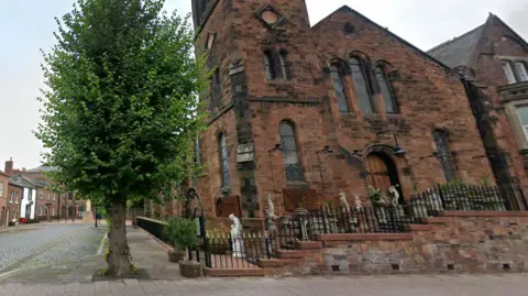 Google A Google Streetview screenshot of the outside of Michelangelo in Carlisle. The red sandstone building has a pitched roof and church-like windows, with statues dotted around the external area, up the stairs at the front of the building