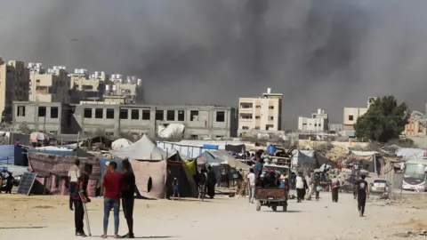 Reuters: Smoke rises after an Israeli attack on a residential building in Khan Younis in the Gaza Strip. In the foreground, tents are set up and people walk around.