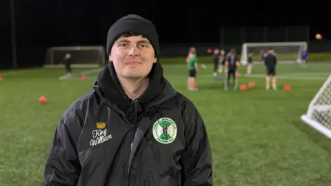 Tyler stands smiling at a training session wearing his black Tunley Athletic-branded coat and black beanie hat