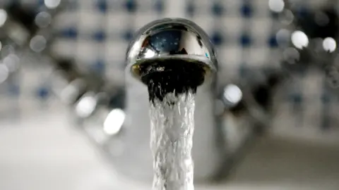 An extreme close up of as silver water tap in a home with water coming out. There are blue and white tiles in the background - they are blurred along wiht the hot and cold water taps.