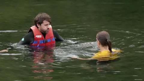 A man with brown hair wearing a red life jacket and black wetsuit visibly exhales while floating in a lake. He is looking at a woman with a ponytail wearing a yellow shirt who is also in the water. The water looks green.