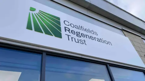 A white sign on a brick building with green writing on which says 'Coalfields Regeneration Trust". There are windows below.
