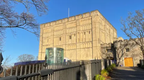 Shaun Whitmore/BBC A Norman castle stands on a mound with clear blue skies above it and trees and metal railings in front of it