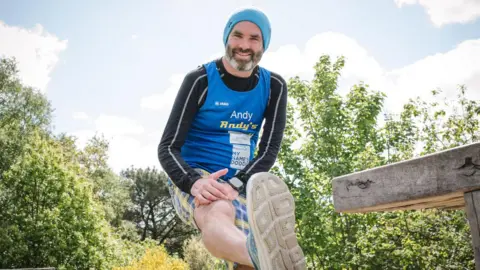 Andy Vaughton is wearing running gear outside, and is stretching his leg.