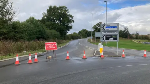 Exit from roundabout coned off, with red "emergency road closure" sign. There is also a roadsign to Kilsby, Ashby St Ledgers and DIRFT. There are trees on one side of the road and grass on the other.