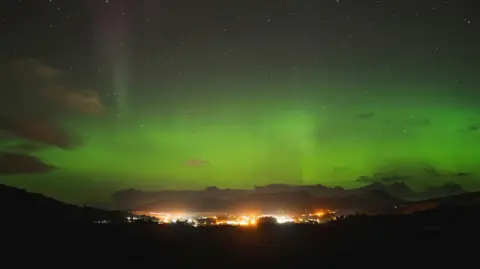 The aurora, also known as the Northern Lights, glows green above a brightly lit community in the Isle of Skye.