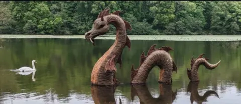 Colin Knox Large Loch Ness monster sculpture rising from calm water in a lake, with a white swan swimming nearby and dense green trees lining the far shore.