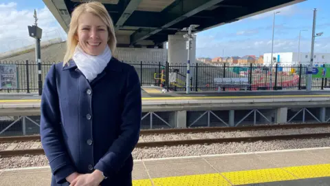 Olivia Perkins is standing on a platform underneath a road bridge. She is wearing a blue suit and a white scarf and has blonde, shoulder length hair.