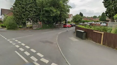 A Google street view image of the entrance to a road from a T-junction. It is a residential street with houses and fences on either side, with a large tree to the left. Cars are parked along the side of the road in the distance.