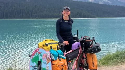 Naomi Hancocks stands in front of blue water with her pink bike which is laden with all the equipment she needs to travel solo. 