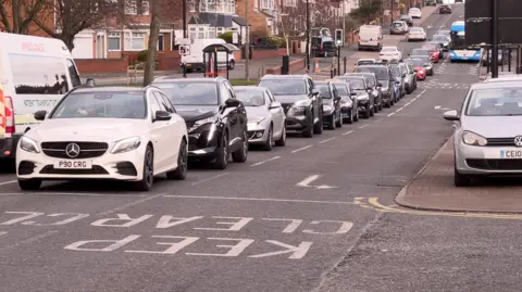 A row of cars waiting for a junction to be clear of other vehicles, while one of the carriageways, currently not a dedicated bus lane, is occupied by a blue and white bus.
