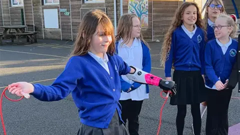 Zoey is in her blue school uniform and is smiling. She has brown hair and a fringe. Part of he bionic arm can be seen as the sleeve of her cardigan has been pushed up. The bionic arm has a white casing and black outlining. She is holding a pink skipping rope on a patterned playground with five other children in the background