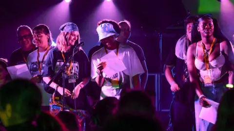 A mixture of men and women are standing up on stage, under a purple sportlight, some stand behind a microphone, they're holding pieces of paper and either looking out at the audience or smiling as they read from the paper. 