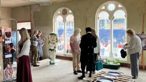 People standing inside a building with large ornate windows. They are looking at a mix of artwork, some is displayed on stands, some on the floor