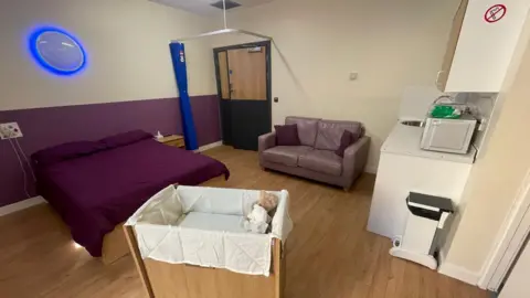 The photograph shows a room with a purple double bed, a sofa and a little kitchen. In the foreground is a cold cot, with two teddy bears inside. The cold cot enables parents to stay longer with their children after they have died.