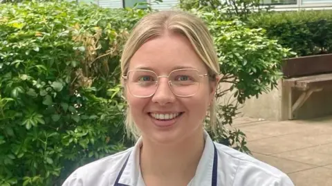 Becky Sheppard is sitting on a bench in front of a bush. She has blonde hair in a ponytail and is wearing a white hospital uniform. She has clear framed glasses.