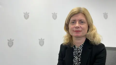 A woman with light, shoulder-length hair with a centre parting and red lipstick. She is wearing a black and white patterned blouse underneath a black blazer. She is sitting on an office-style chair, in front of a white backdrop which features black, Government of Jersey crests. She is staring directly at the camera, to the right of the image.