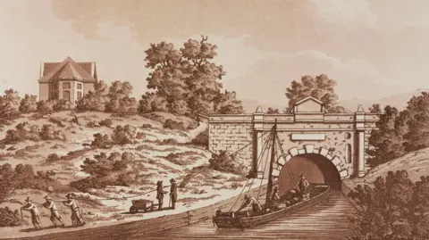 SSPL/Getty Images A historical sepia-toned print, illustrating a barge about to enter the tunnel on the Thames and Severn Canal at Sapperton Hill, Gloucestershire. In the distance, the old Tunnel House sits at the top of a hill. Men stand on the bank, pulling rope or talking to each other.