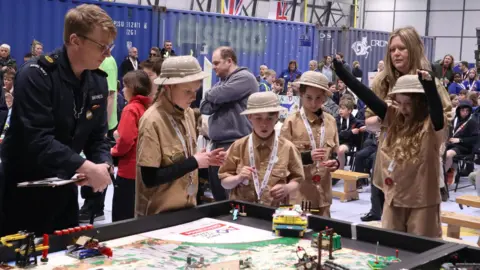 A group of school pupils wearing archaeology outfits – light brown shirts, trousers and hats, stand by a table as their Lego robot completes a series of challenges. A judge from the Royal Navy is watching on. Lots of other scholochildren and adults are standing behind them.