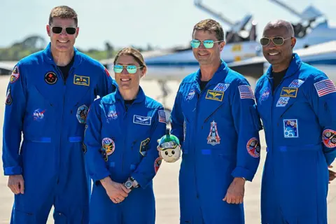 Miguel J. Rodriguez CARRILLO / AFP via Getty Images Artemis crew stand side by side in blue overalls and sunglasses, smiling at the camera 