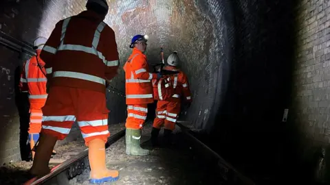 Network Rail Workers in orange protective clothing, helmets and torches around their heads are surveying a dark railway tunnel at Whitehaven.