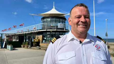 A man wearing a white shirt with naval-style epaulettes smiles as he stands in front of a modern-looking building with a glass tower, canopy and five Union flags flying under a blue sky with the sea in the background.   