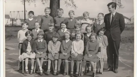 Family A black and white photo of 19 children sitting on three rows of chairs outside for a school photo, with their teacher in a black suit standing next to them.
