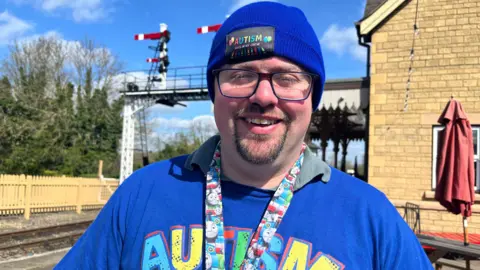 Emma Baugh/BBC James Rollow, a man wearing a blue T-shirt, a blue hat which has AUTISM written on it and a Thomas the Tank Engine-branded lanyard, smiling for the camera. 
