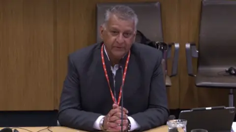 Nilesh Sachdev sat at a desk at a Senedd committee, with a glass of water and a tablet computer in front of him. He is wearing a suit and a white shirt.