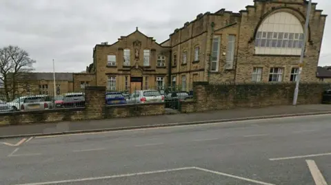 Google The outside of a school. Architecturally, the building probably dates to the early 20th century and is pictured from behind a boundary wall. Some parked cars are visible in the car park.