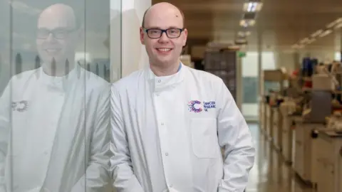 Cancer Research UK Prof Owen Sansom is wearing a Cancer Research UK branded lab coat with his hands in his pockets. He is leaning against a glass panel which is showing his reflection