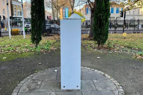 An artwork featuring a comic white cockatiel bird lying on its back on a stone plinth with a park, road and buildings in the background