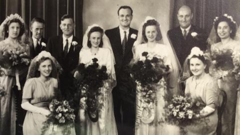 A black and white photograph of Joan and Monica dressed in bridal gowns, veils and holding bouquets and standing besides their husbands and wedding party.