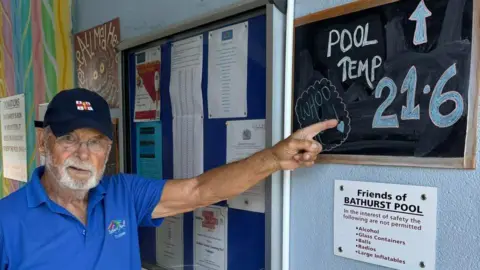 Facebook A photo showing a white man with a beard, he is wearing glasses and a blue RNLI cap. He is stood next to a board which reads pool temp 21.6 degrees. There is a white friends of Bathurst Pool sign underneath