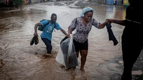 EPA/Shutterstock A man, wearing a blue T-shirt and carrying his shoes, follows behind a woman holding a plastic bag and being helped by another person off-frame as they wade through knee-deep muddy flood water