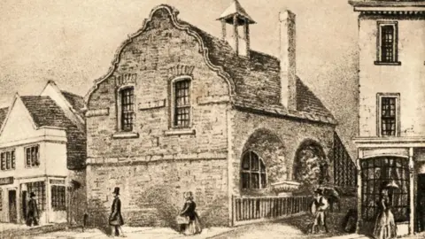 The Norris Museum A line drawing showing the exterior of the Cromwell Museum in Huntingdon in the mid-19th Century. It shows a stone medieval building on a street with people walking in front and there are shops on either side. 
