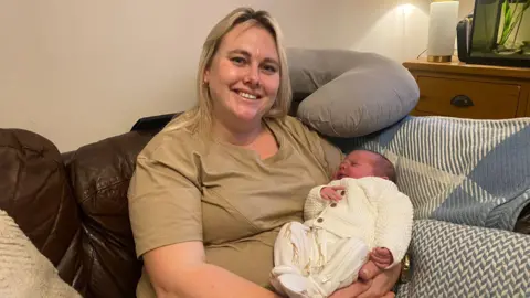 A blonde woman wearing a light-brown coloured t-shirt is smiling and cradling her new born baby in her arms. The baby is dressed in a white romper with giraffes on. They are sat on a sofa and there is a neck pillow propped up behind her.
