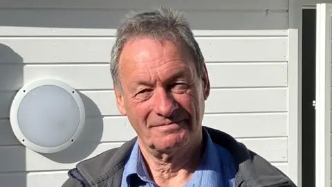 A man with grey hair, in a blue shirt and dark jacket standing in front of a white weather-board building with a light to his right. 